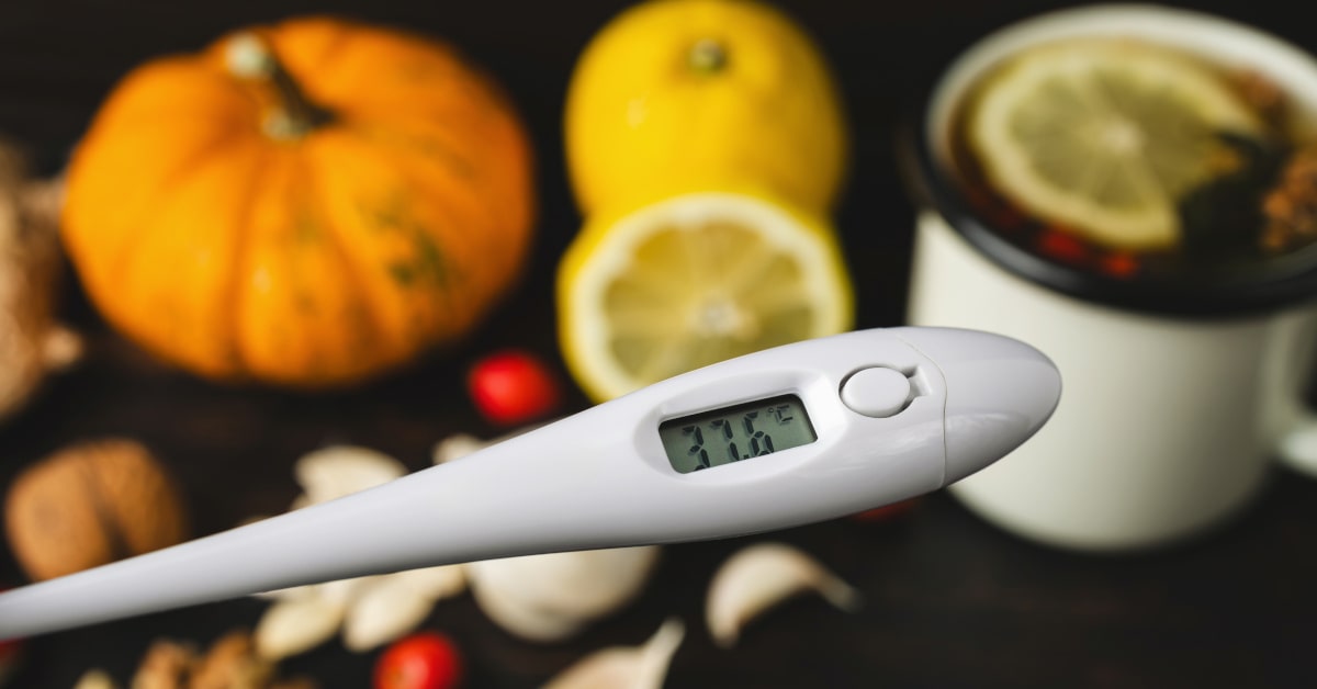Termometro digitale che indica 37.6 &deg;C e sullo sfondo rimedi naturali e tisana calda